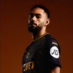 Portrait of a man with short hair and a beard, wearing a dark athletic jersey with a white circular patch on the sleeve, against an orange background.