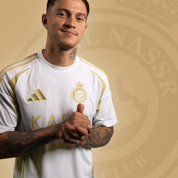 Portrait of a young man in a white and gold soccer jersey with a crest, arms folded, against a gold emblem backdrop