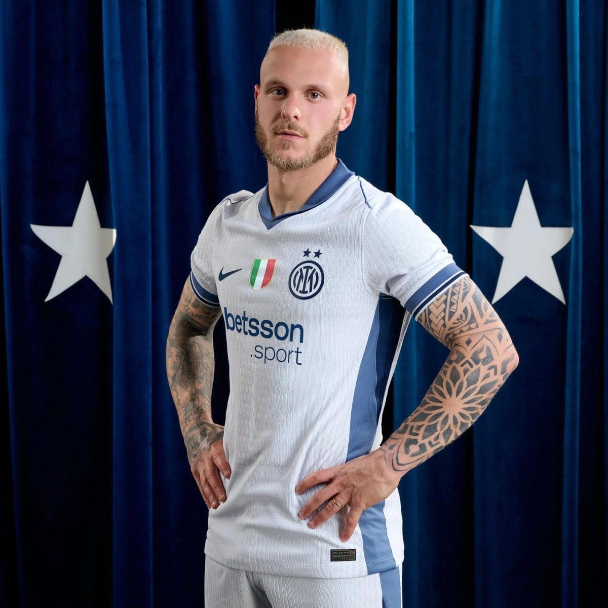 Footballer wearing an Inter Milan jersey posing with hands on hips against a blue curtain backdrop with white stars behind him.