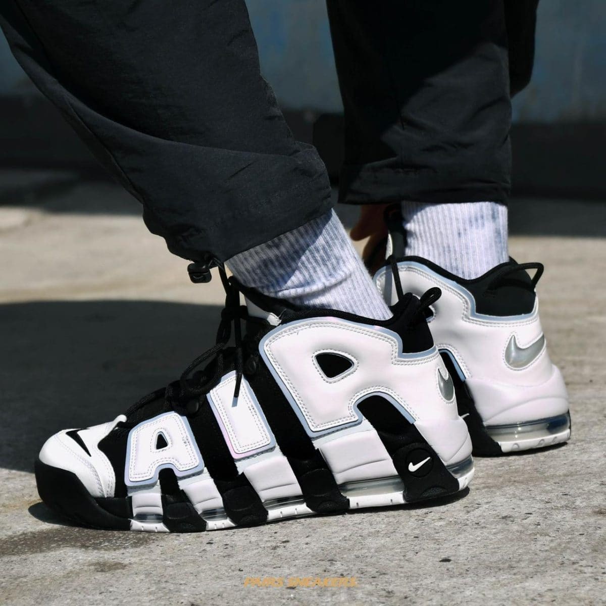 Close-up of white and black Nike Air More Uptempo sneakers worn with gray socks and black pants on a concrete surface.
