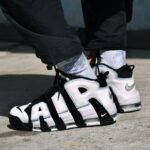 Close-up of white and black Nike Air More Uptempo sneakers worn with gray socks and black pants on a concrete surface.