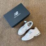 White New Balance running shoes placed beside a dark blue New Balance shoebox on a beige carpet floor.