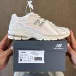 Light pink and white New Balance 1906RLI sneaker with tag attached, resting on a dark shoe box labeled 'New Balance' and size details visible.