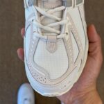 Close-up of a white/cream athletic sneaker being held, showing the laces and toe box with visible stitching and a small 'N' logo plate near the laces.