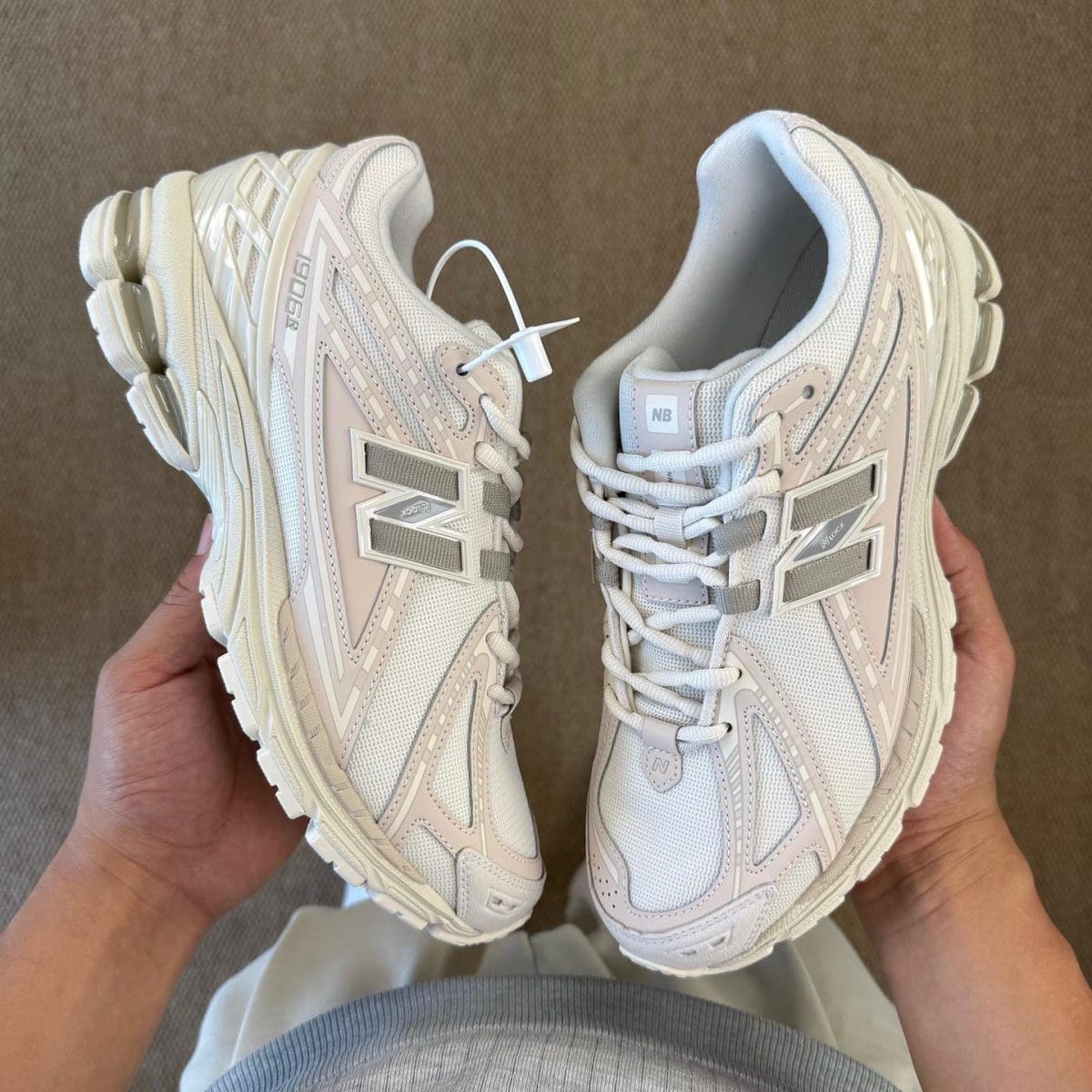 Pair of white/cream New Balance sneakers being held, showing side and toe details with prominent 'N' logo.