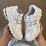 Pair of white/cream New Balance sneakers being held, showing side and toe details with prominent 'N' logo.