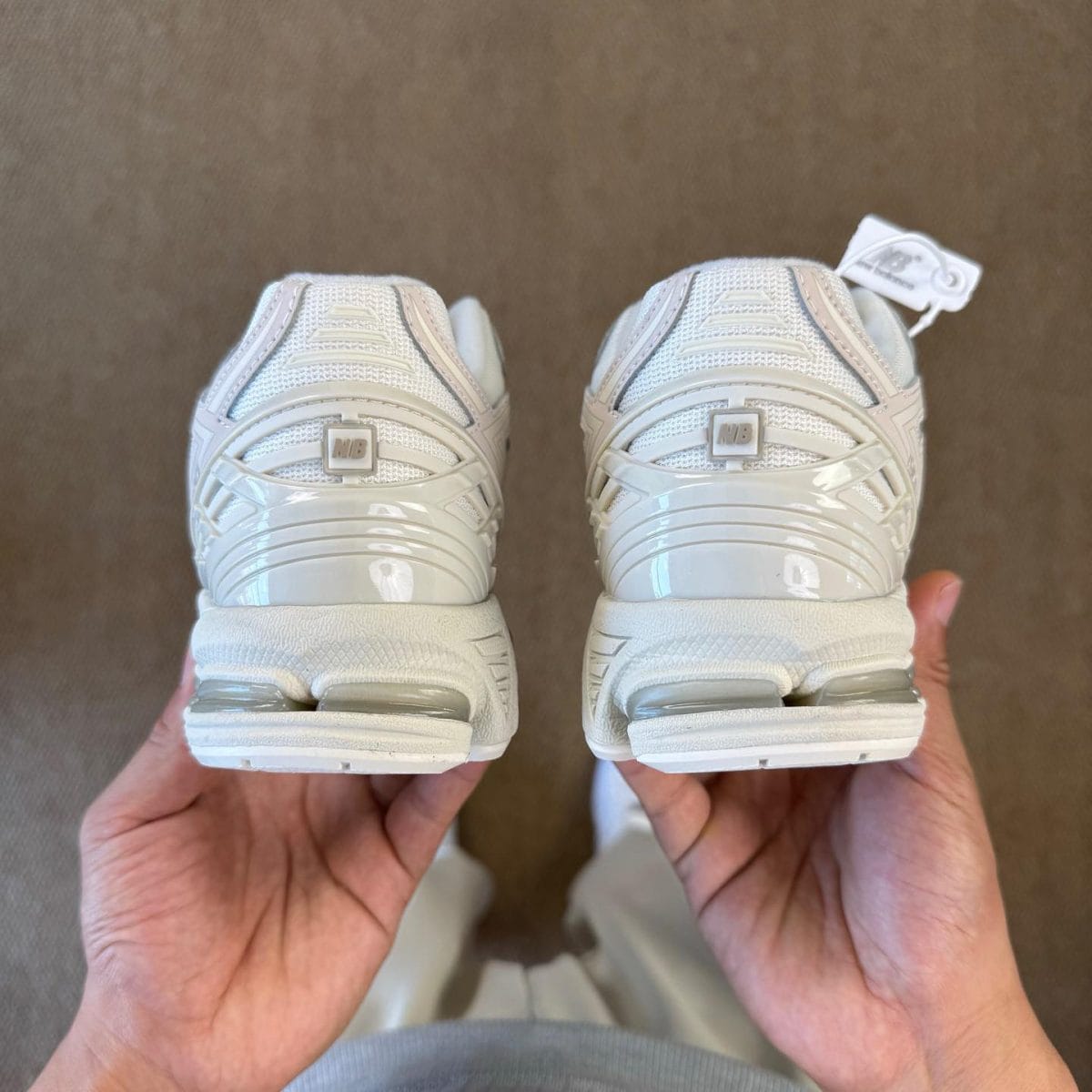 Two white sneakers held from behind, showing textured uppers, chunky heel counters, and an NB logo on each heel.