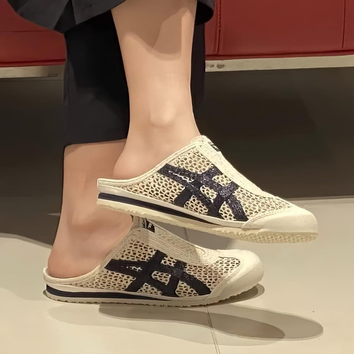 Product photo showing a white mesh slip-on sneaker with dark blue crisscross stripes, worn on a mannequin leg from the side.
