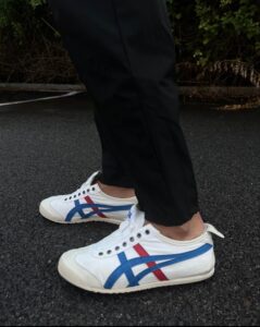 Close-up of white sneakers with blue and red stripes worn with black pants, standing on dark asphalt surface in an outdoor setting.