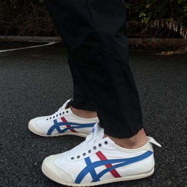 Close-up of white sneakers with blue and red stripes worn with black pants, standing on dark asphalt surface in an outdoor setting.
