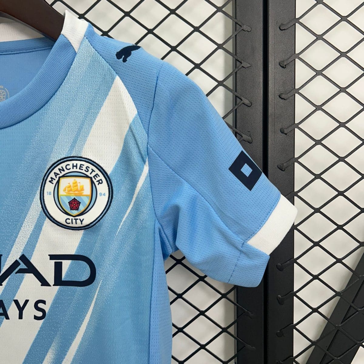 Light blue Manchester City soccer jersey on a hanger, with the club crest visible, against a metal chain-link fence background.