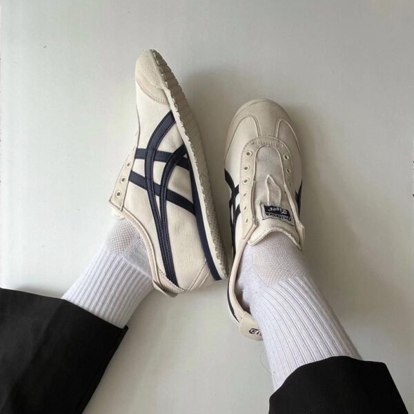 Two white sneakers with dark blue stripes and white socks resting on a light floor, feet crossed at the ankles.
