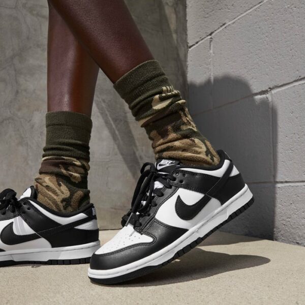 Person wearing black-and-white Nike sneakers with camouflage socks standing on a concrete step next to a gray wall.
