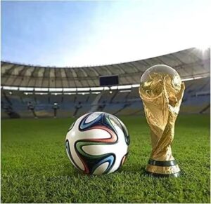 Soccer ball and FIFA World Cup trophy on a green stadium field with empty stands in the background.