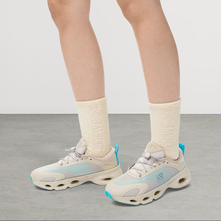 51d5d2df-8e76-4ff8-a38b-bf7884a4b444 Pair of light beige and aqua athletic sneakers with cream socks, shown on a gray floor from the calves down for a fashion/footwear look.