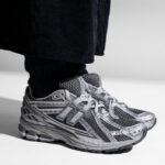 51d938f8-dfde-40e6-9170-86981382ba5f | UAE SPORT STORE Gray athletic sneaker with a large N-shaped logo on the side, worn with black pants by a person standing.