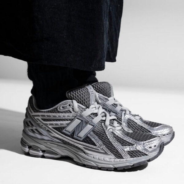 Gray athletic sneaker with a large N-shaped logo on the side, worn with black pants by a person standing.