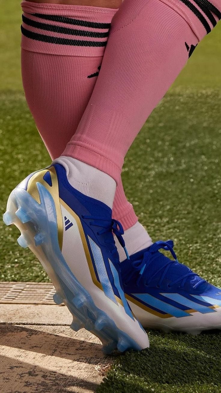 51f8430fbf1b5d19bacd9d858c38430d | UAE SPORT STORE Close-up of a blue and white soccer cleat with gold accents on a player's pink knee-high sock, on artificial turf.
