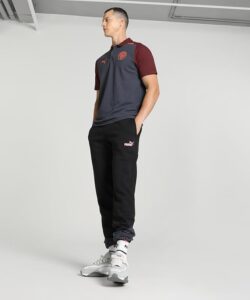 Man modeling athleticwear: dark gray and maroon polo, black joggers with Puma logo, white sneakers, hands in pockets in a neutral studio setting