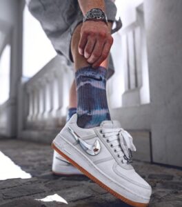 White low-top Nike sneakers with a gum sole, on a person wearing blue tie-dye socks, captured mid-step on a cobbled surface.