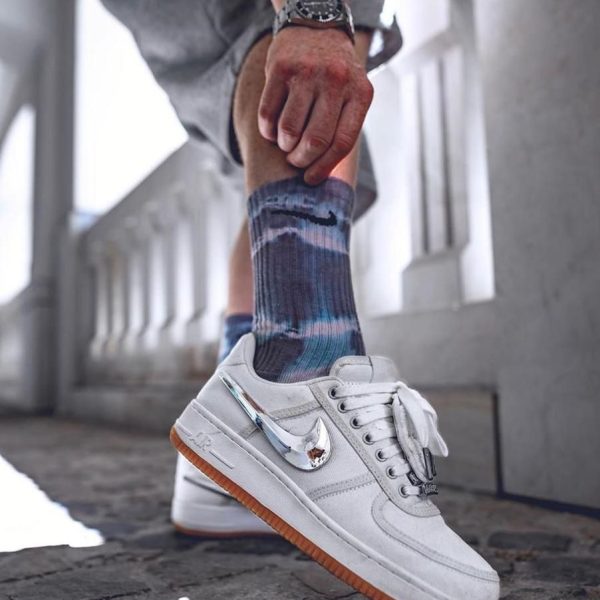 White low-top Nike sneakers with a gum sole, on a person wearing blue tie-dye socks, captured mid-step on a cobbled surface.