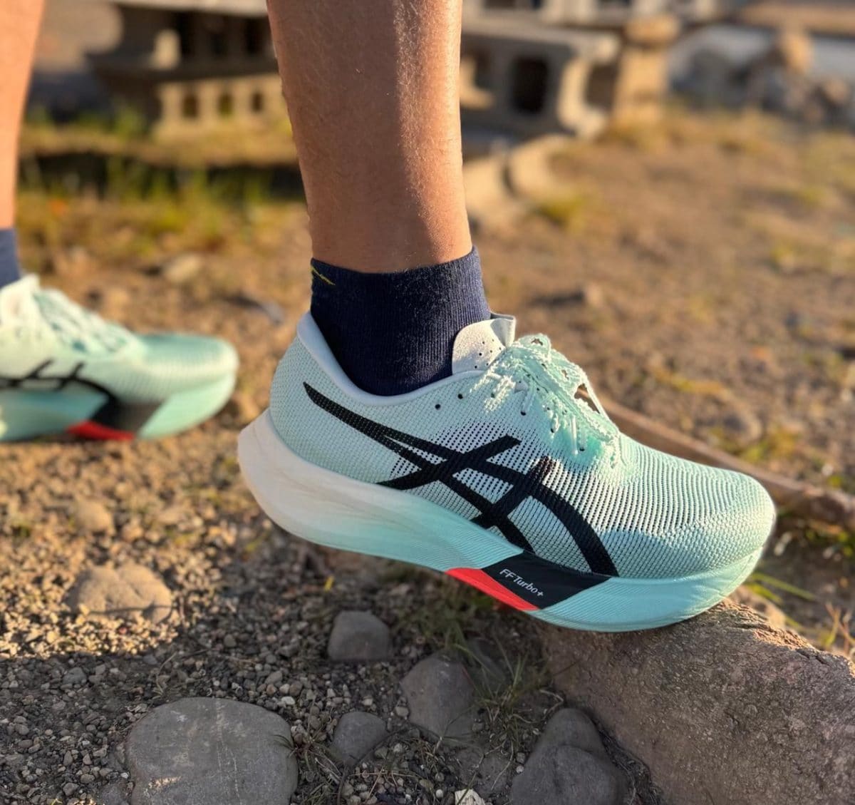Teal running shoe on a rocky trail, close-up of laces and white sole, with a dark sock visible above the shoe.