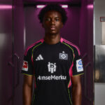 Young Black male soccer player in a black jersey with pink trim, standing in a purple-lit hallway.