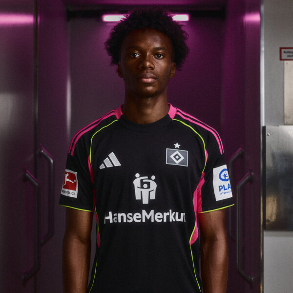 Young Black male soccer player in a black jersey with pink trim, standing in a purple-lit hallway.