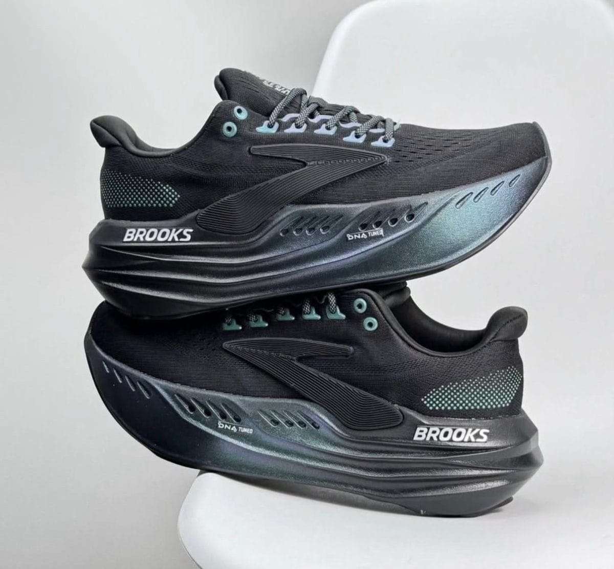 Two black Brooks running shoes stacked on a white chair, with teal eyelets and a white BROOKS logo on the midsole.