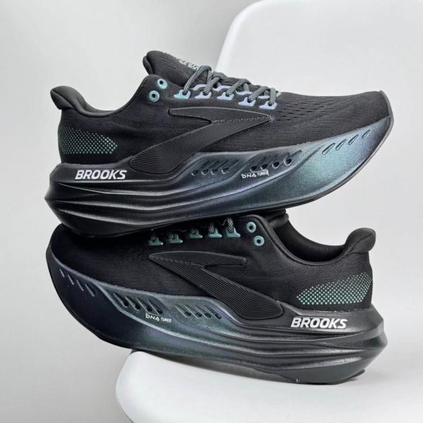 Two black Brooks running shoes stacked on a white chair, with teal eyelets and a white BROOKS logo on the midsole.