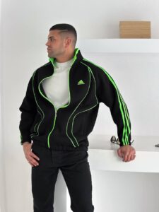 Man wearing a black Adidas tracksuit with neon green piping, standing sideways in a bright white room with a shelf in the background.