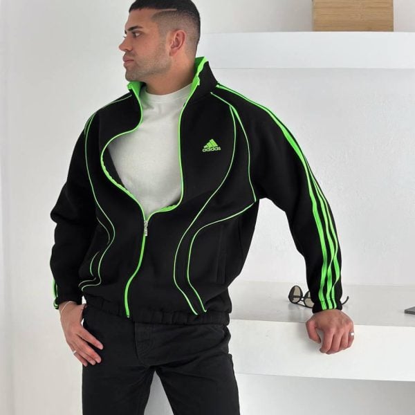 Man wearing a black Adidas tracksuit with neon green piping, standing sideways in a bright white room with a shelf in the background.