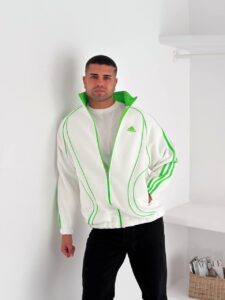 Man wearing a white Adidas track jacket with neon green piping, standing against a white wall.