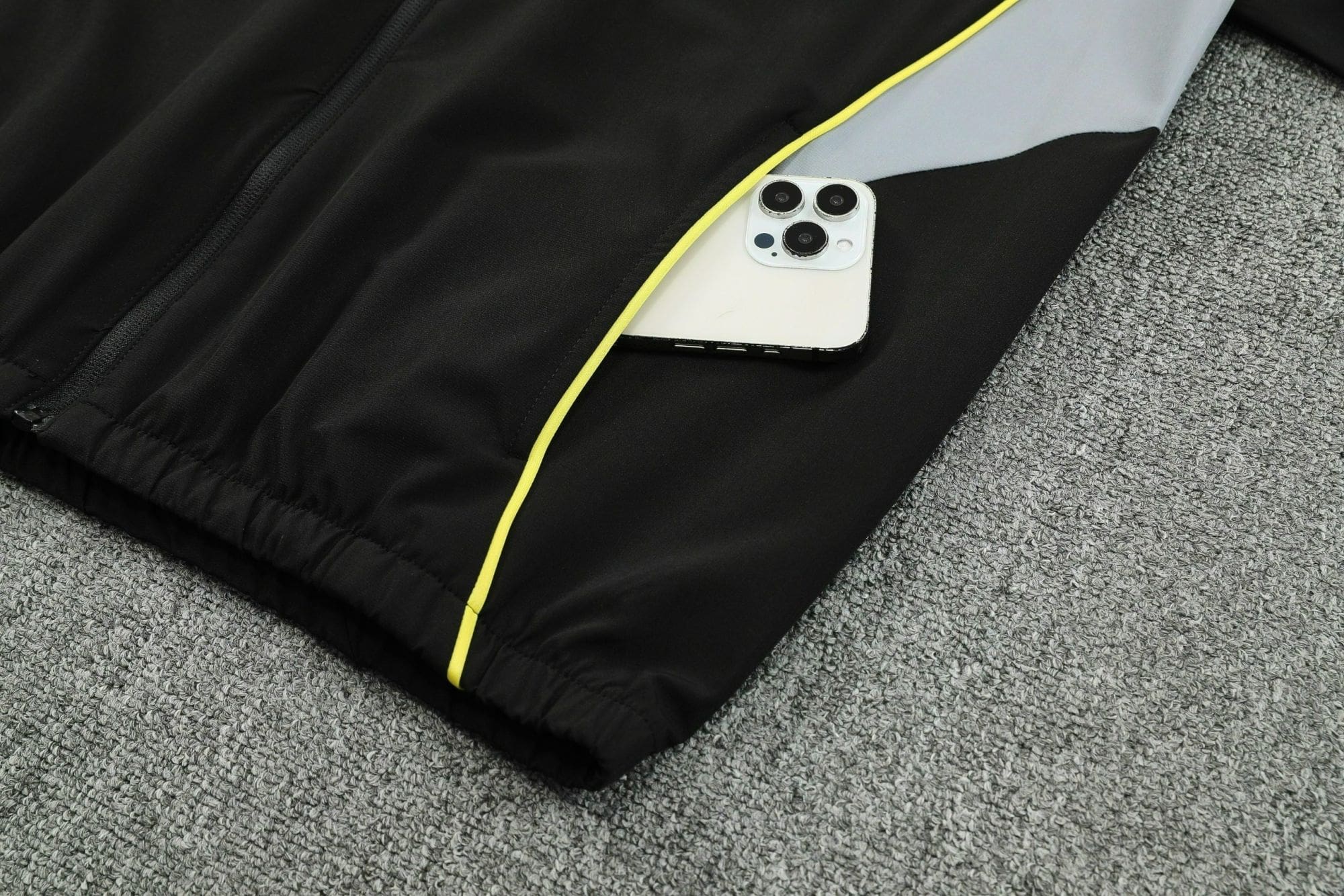 5328bfb3 | UAE SPORT STORE Close-up of a black jacket with neon yellow zipper trim, partially covering a white iPhone resting on a gray carpet.