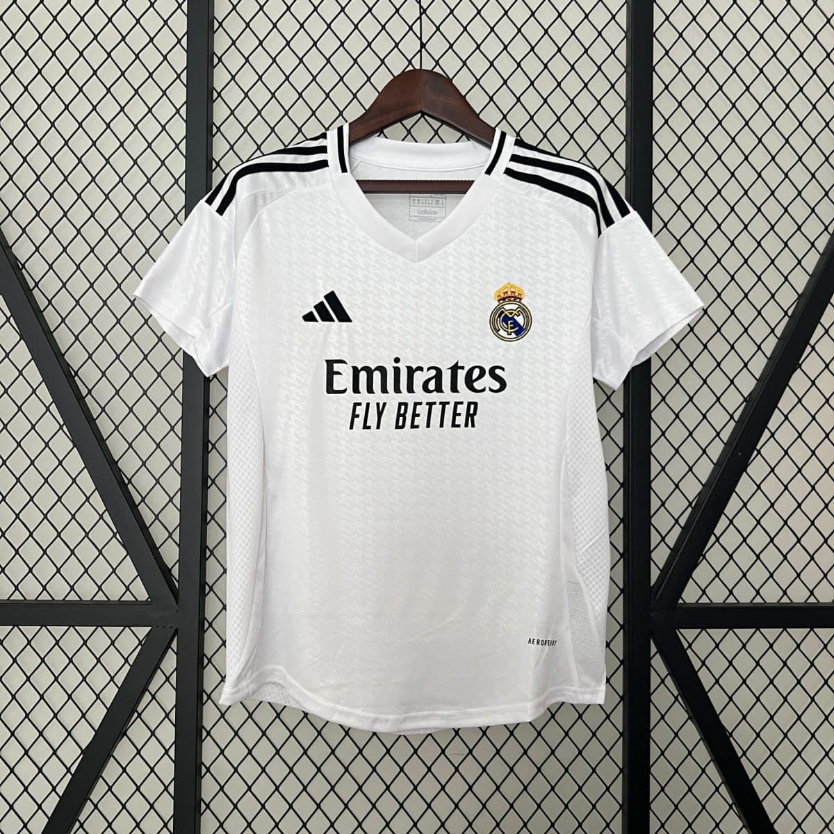 White Real Madrid jersey on a hanger against a diamond-pattern metal grid, showing the crest, Adidas logo, and Emirates sponsor text.