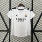 White Real Madrid jersey on a hanger against a diamond-pattern metal grid, showing the crest, Adidas logo, and Emirates sponsor text.