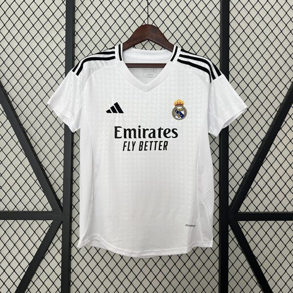 White Real Madrid jersey on a hanger against a diamond-pattern metal grid, showing the crest, Adidas logo, and Emirates sponsor text.