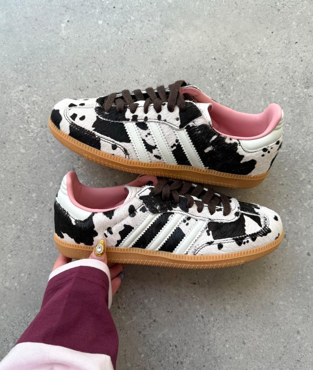 5399a81f-c8fe-4056-9d15-ac8ae75c7069 | UAE SPORT STORE Pair of cow-print sneakers with white stripes, brown laces, pink interior, and gum soles laid on a concrete surface.