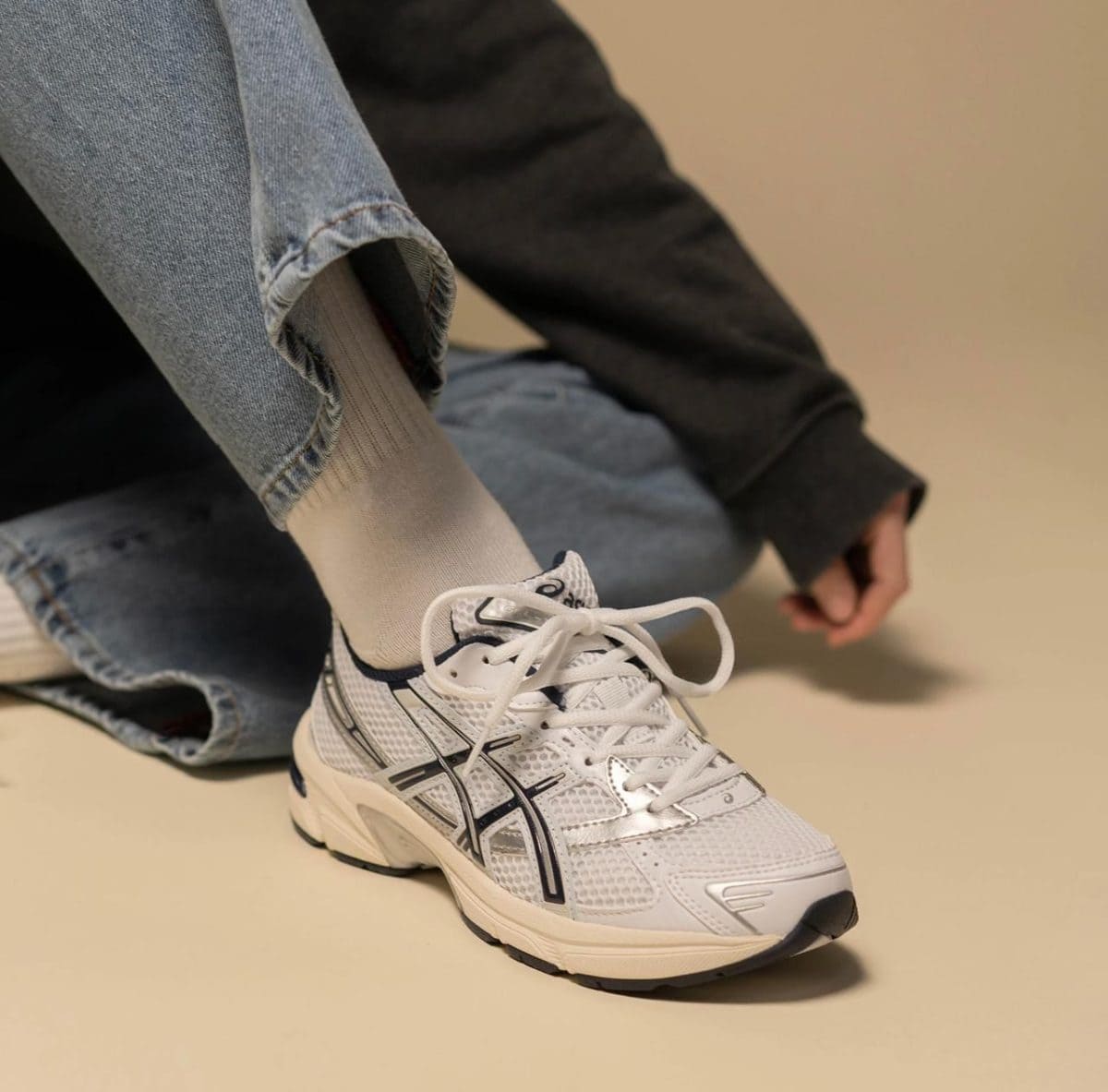 Close-up of a white athletic sneaker with laces tied, worn with light beige socks and cuffed jeans, sitting on a beige floor