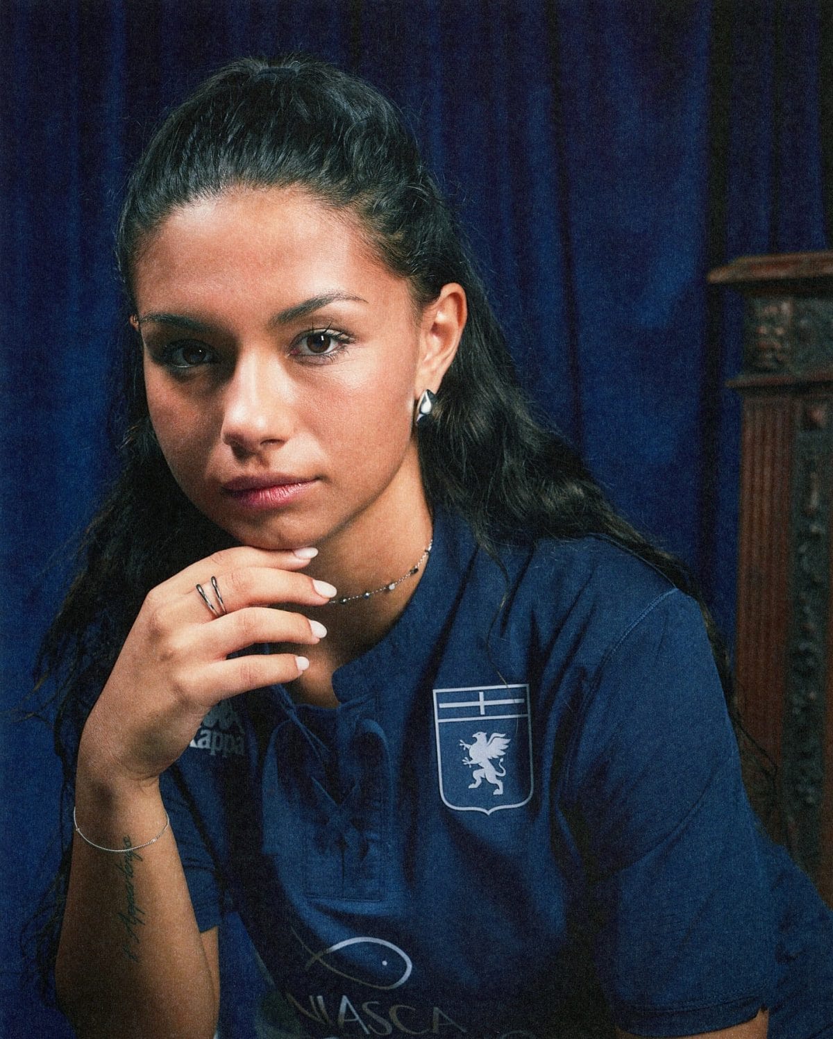 Portrait of a young woman in a blue uniform with a crest, resting her chin on clasped hands against a dark blue curtain backdrop, looking at the camera.