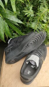 Pair of black sneakers with thick, circular-sole tread resting on a tan surface beside green leafy plants.