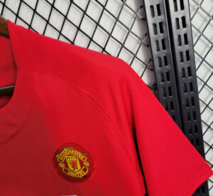 Red Manchester United football jersey with club crest visible on chest, hanging on a metal rack.