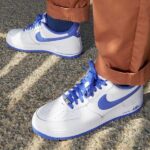 Pair of white Nike Air Force 1 sneakers with blue swooshes and laces on a textured pavement, cropped pants in brown/orange color nearby.