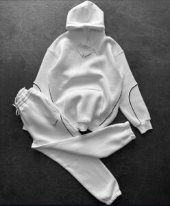 White Nike hoodie and matching joggers laid flat on a dark textured surface, small Nike swoosh on chest and on pants' leg seams.