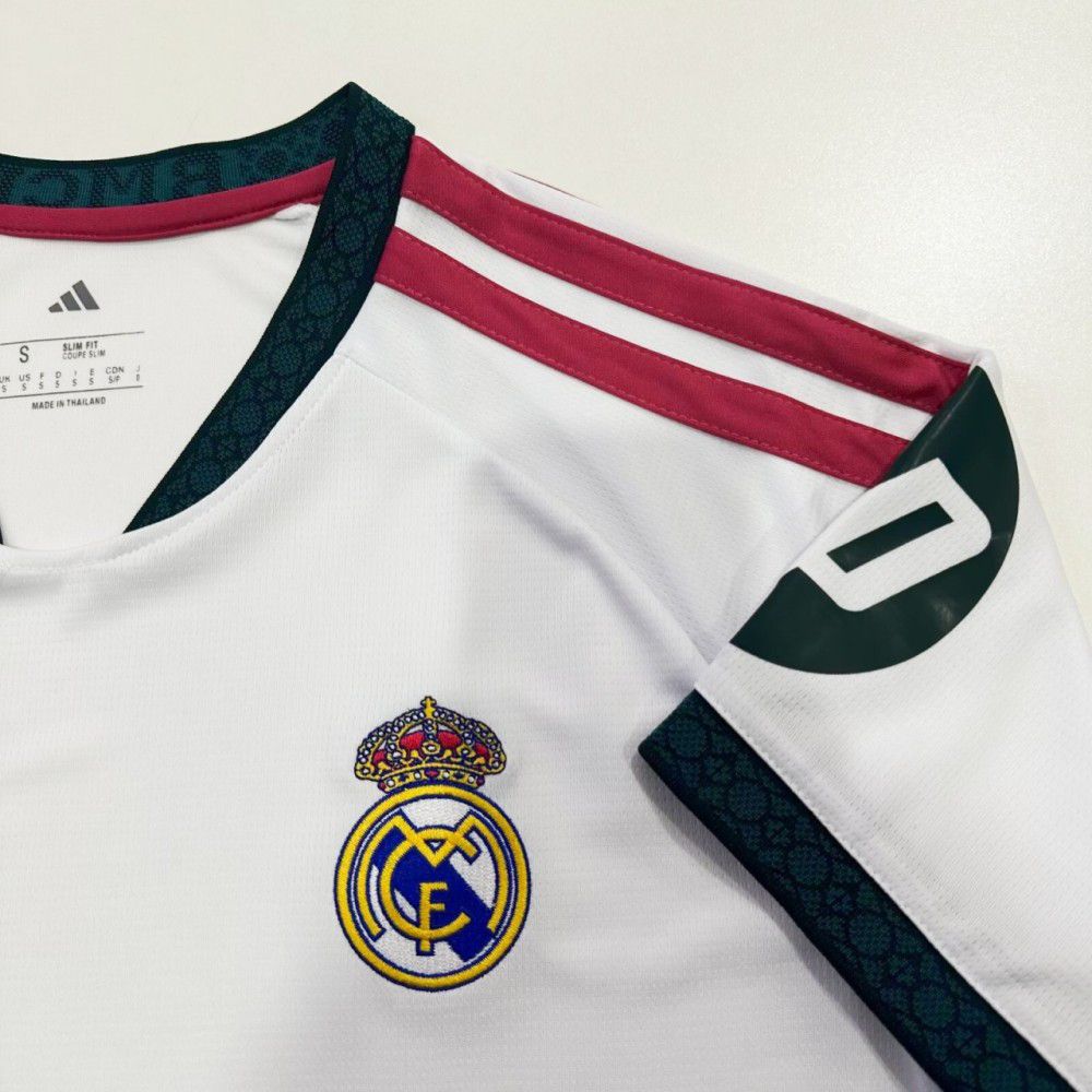 558c8eb4-dc16-4b9e-9355-3a09f6ecae0b | UAE SPORT STORE White Real Madrid football jersey close-up showing the crest, maroon shoulder stripes, and dark patterned trim on the sleeves and collar beside the neck tag.