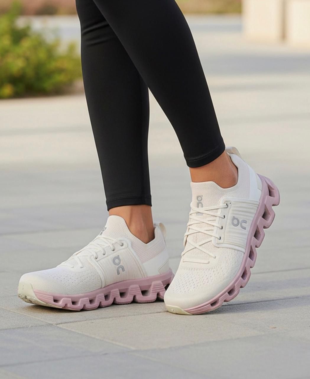 5594e5fd-dbd3-4e47-84d3-1d91e613db33 | UAE SPORT STORE White athletic sneakers with a pink segmented sole on a gray sidewalk, worn with black leggings.