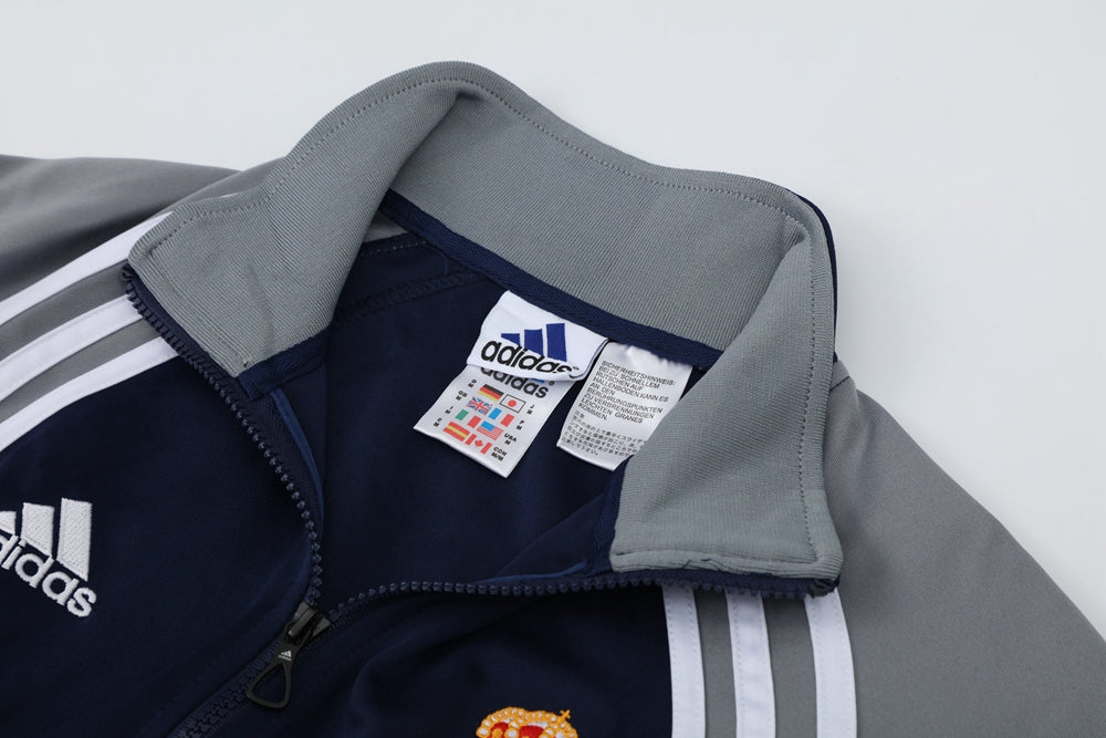 55a106e1 | UAE SPORT STORE Close-up of a navy Adidas track jacket with a gray folded collar, white stripes on the sleeve, and a partially unzipped front zipper.