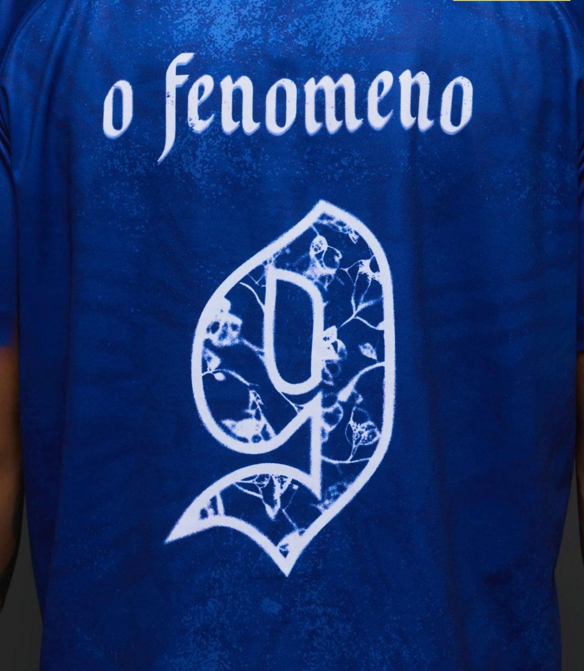 Close-up of a blue T-shirt featuring white text that reads 'o fenomeno' and a large decorative number 9 with a vine pattern.