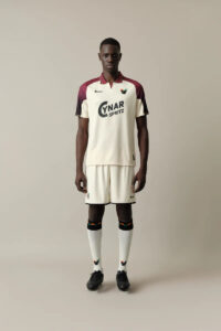 Male soccer player model standing in a cream and maroon kit with sponsor text on the chest.