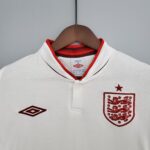 56181139_fe4dcc91-f0cf-4a1e-aa43-dfe7a876323a | UAE SPORT STORE White polo shirt on a wooden hanger with red trim at the neckline and a red crest on the chest, hanging against a gray background.